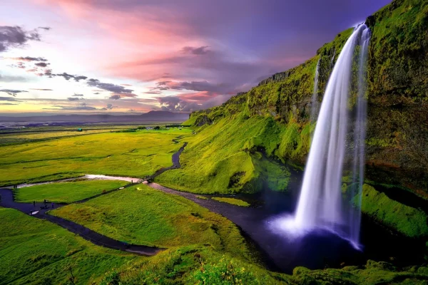 large seljalandsfoss waterfall surrounded by green fields and small river seen during sunset in iceland