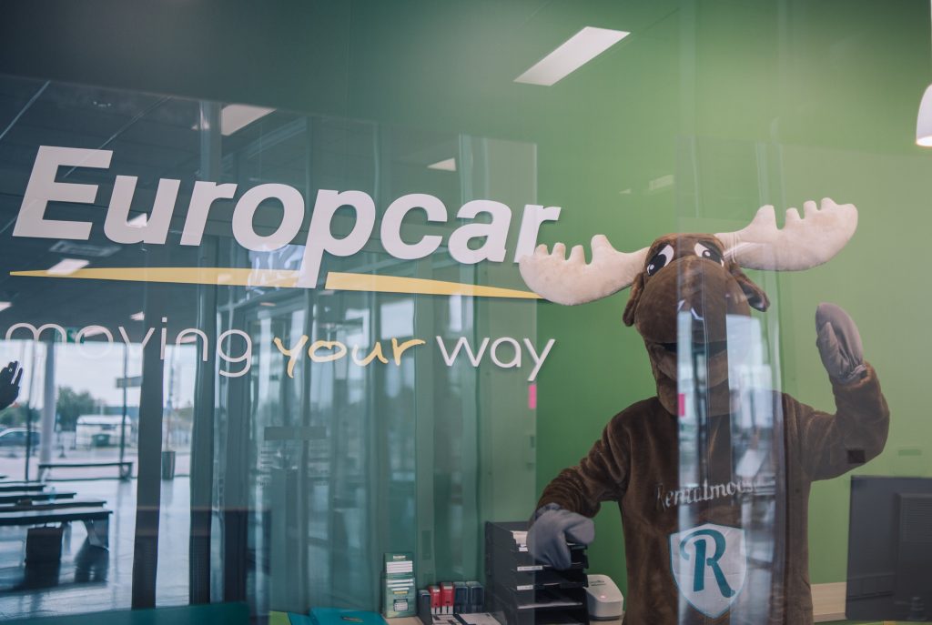 happy rental moose mascot standing next to Europcar car rental banner, behind a glass panel at Europcar counter.