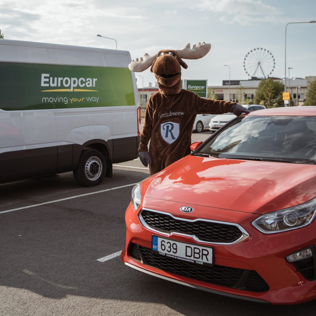 Rental Moose mascot posing next to red Kia rental car. White Europcar van in the background, Europcar banner and ferris wheel in the background