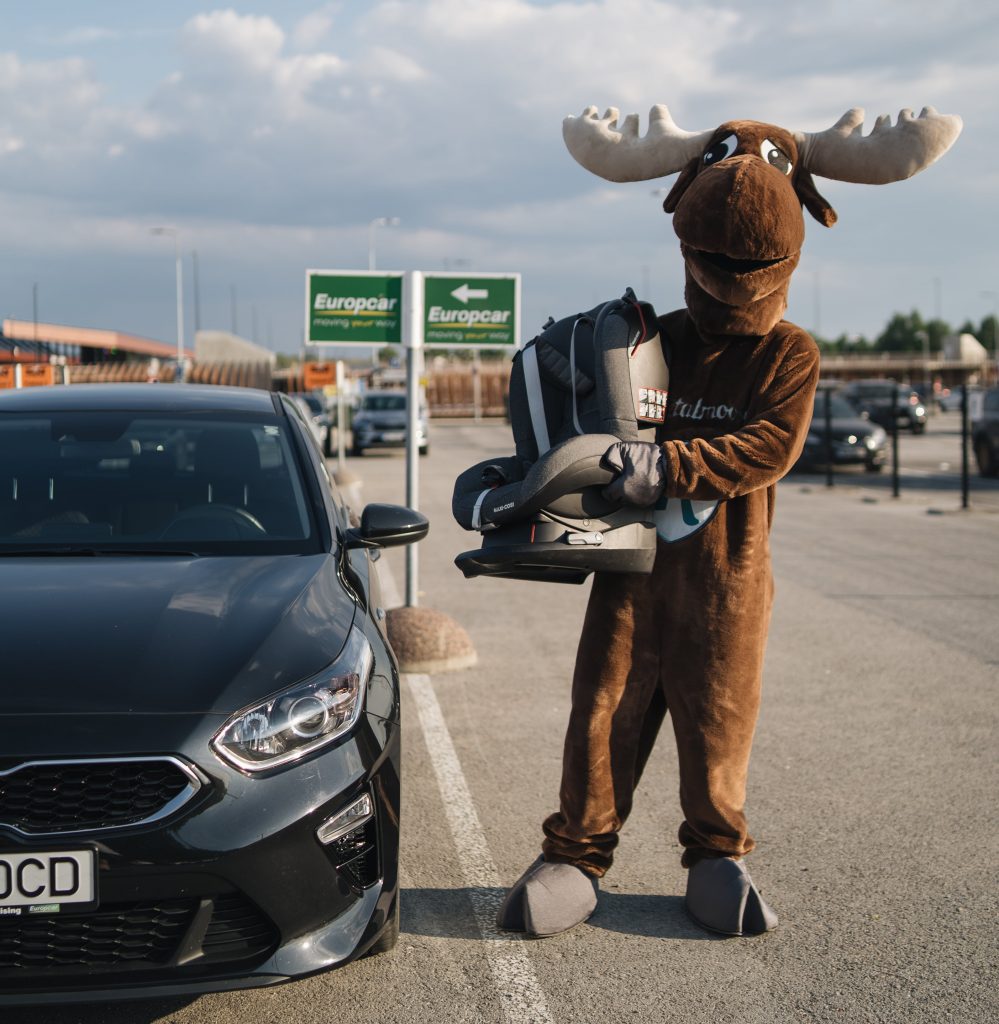 Moose holding child seat next to Kia rental car from Europcar, large carpark during sunset.