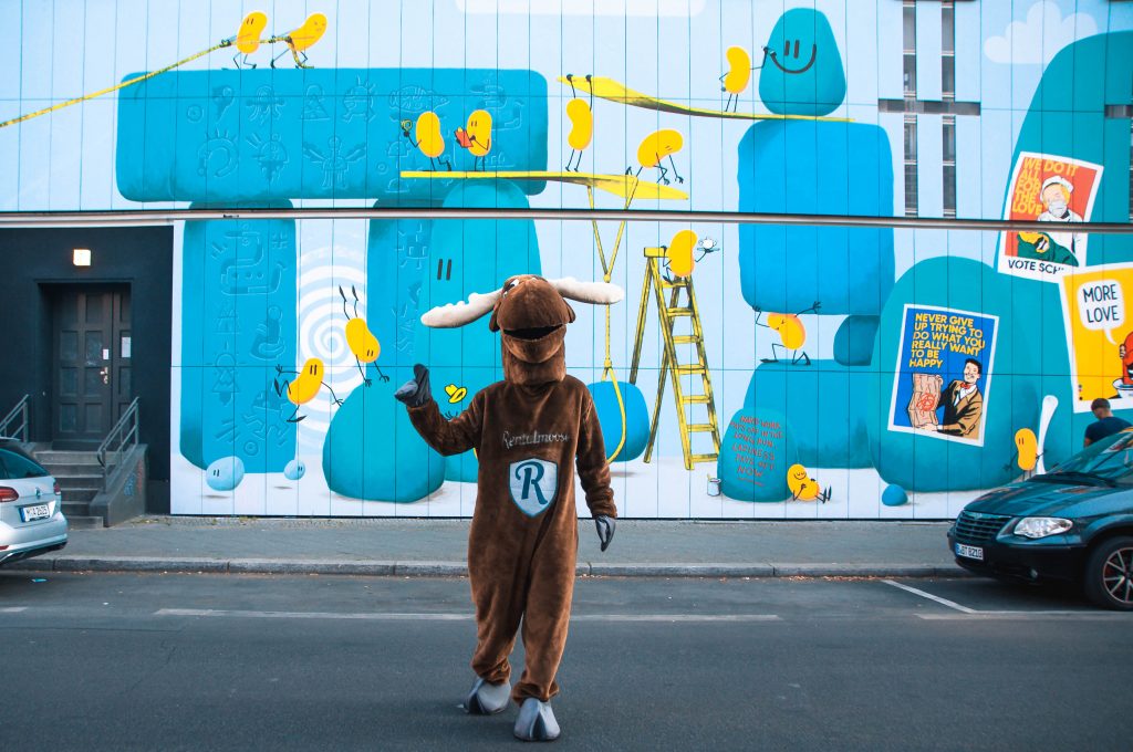 This blog post will cover everything you need to know about security deposits when renting a car. Rental Moose mascot crossing a road in Berlin, colorful blue and yellow mural in the background. 