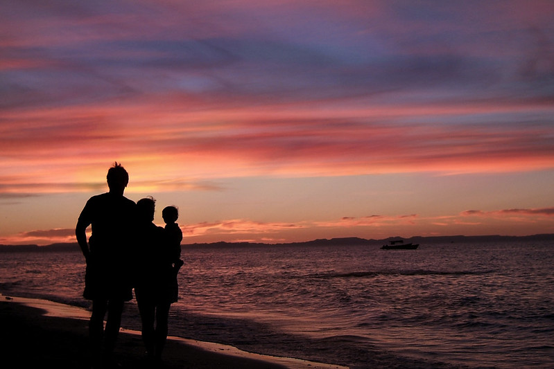 A family of three watching the sun set on a sandy beach in La Paz, Mexico. Some say Baja California is home to some of the most wonderful sunsets on the planet, and this vibrant photograph proves it.