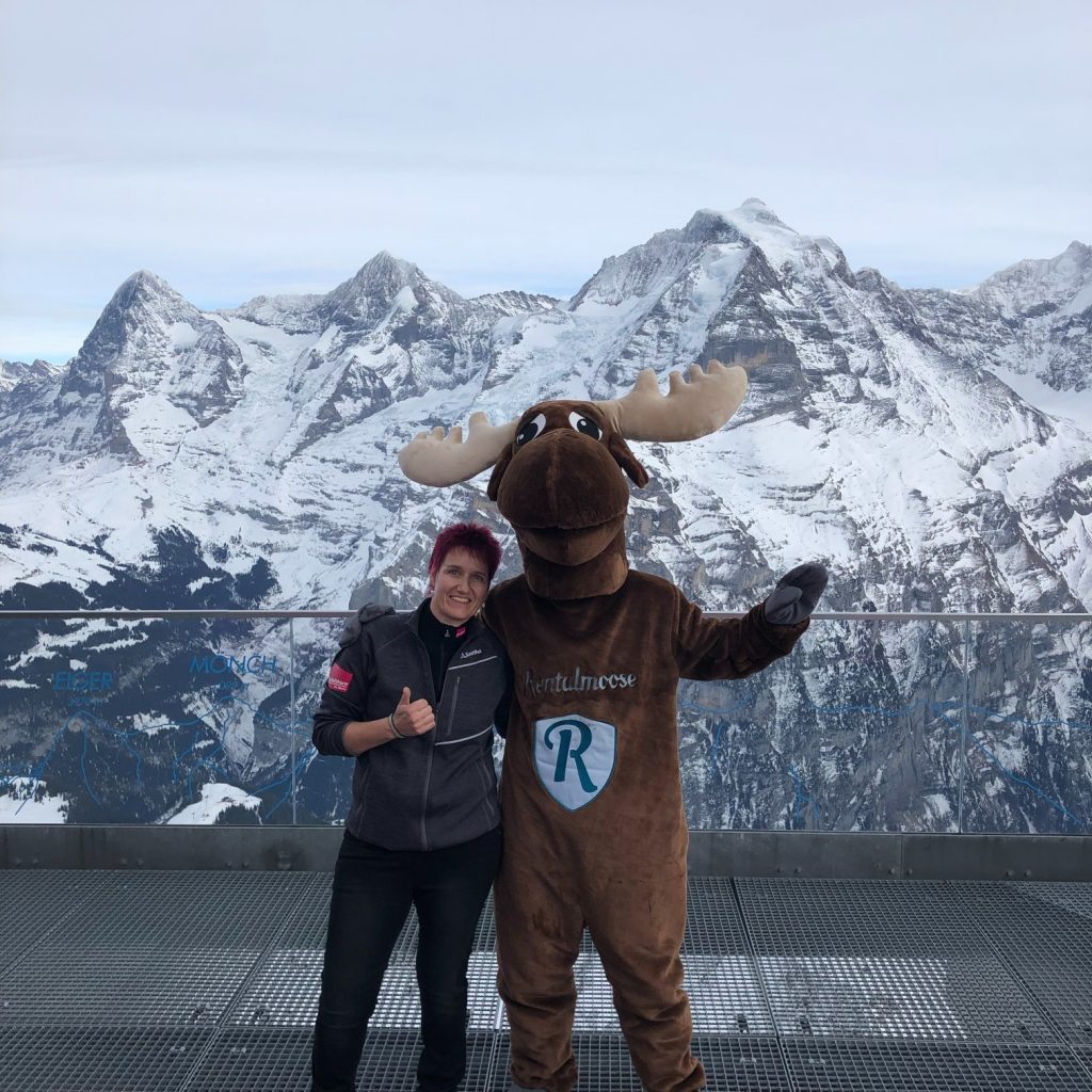 Rental Moose Mascot posing in front of swiss alps panoramic view with local employee. Photo at the summit, winter in Switzerland.