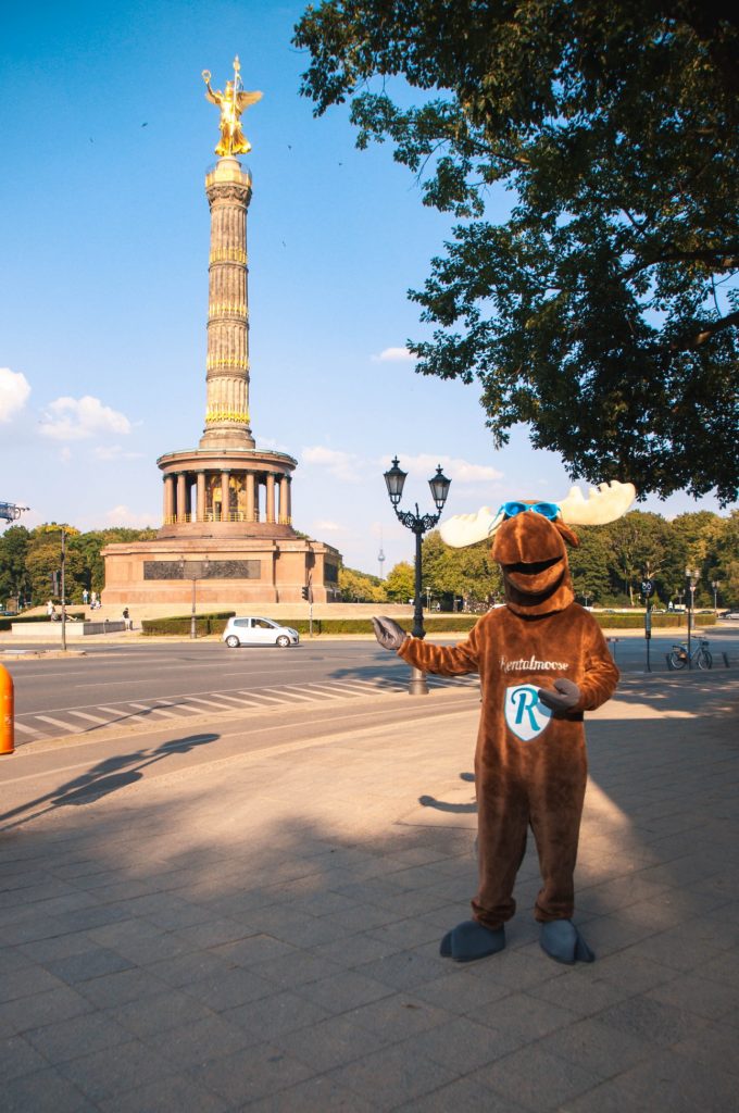 Rental Moose mascot posing in front of Victory Column, a landmark in Berlin. See the best places to visit in Berlin with our curated berlin travel guide