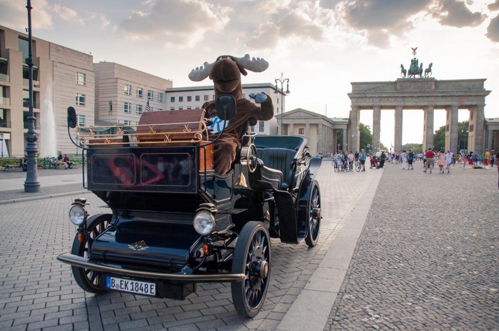 Rental Moose mascot sitting in oldtimer vintage car posing in front of Brandenburg Gate, one of the best things to do in Berlin. See it during our Poland road trip itinerary