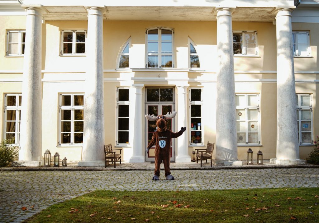 Rental Moose mascot posing with the grand entrance to Kau Manor in Estonia. See the best manors in estonia on our road trip itinerary, the best things to do in estonia