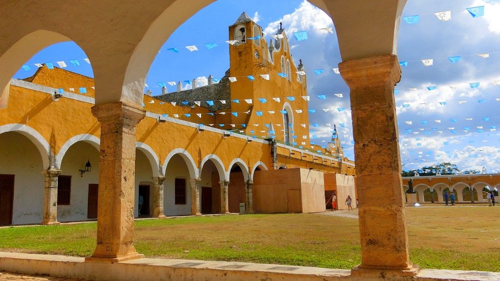 beautiful yellow church and courtyard with blue and white flags over green yard in the town of izamal. A must-see town on a mexico road trip.