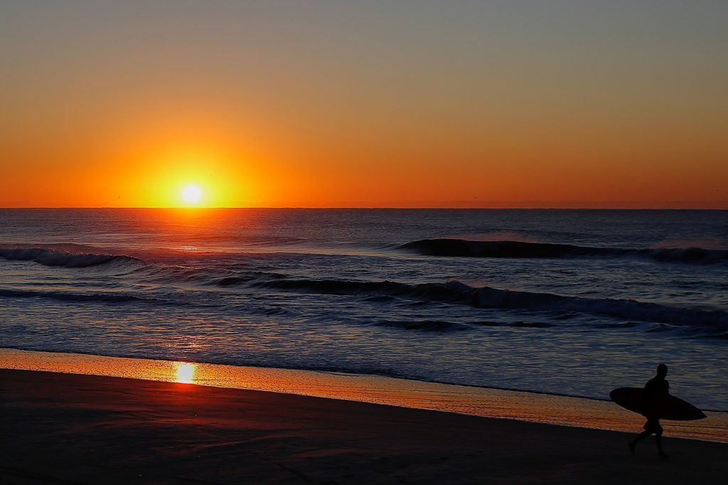 surfer strolling down a sandy beach at sunset, orange sky and calm waves on the ocean. Beautiful evening in Campeche, Yucatan Peninsula, Mexico.