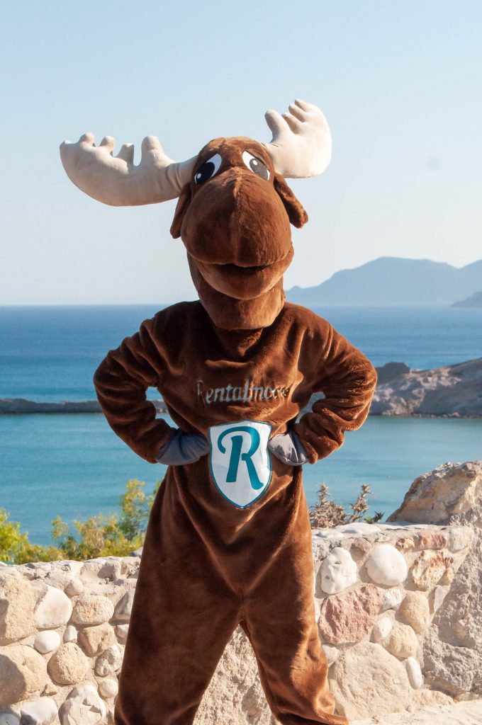 Rental Moose mascot posing at a viewpoint overlooking a beautiful sandy beach in Kos, Greece. Check out our curated Greek Island itineraries.