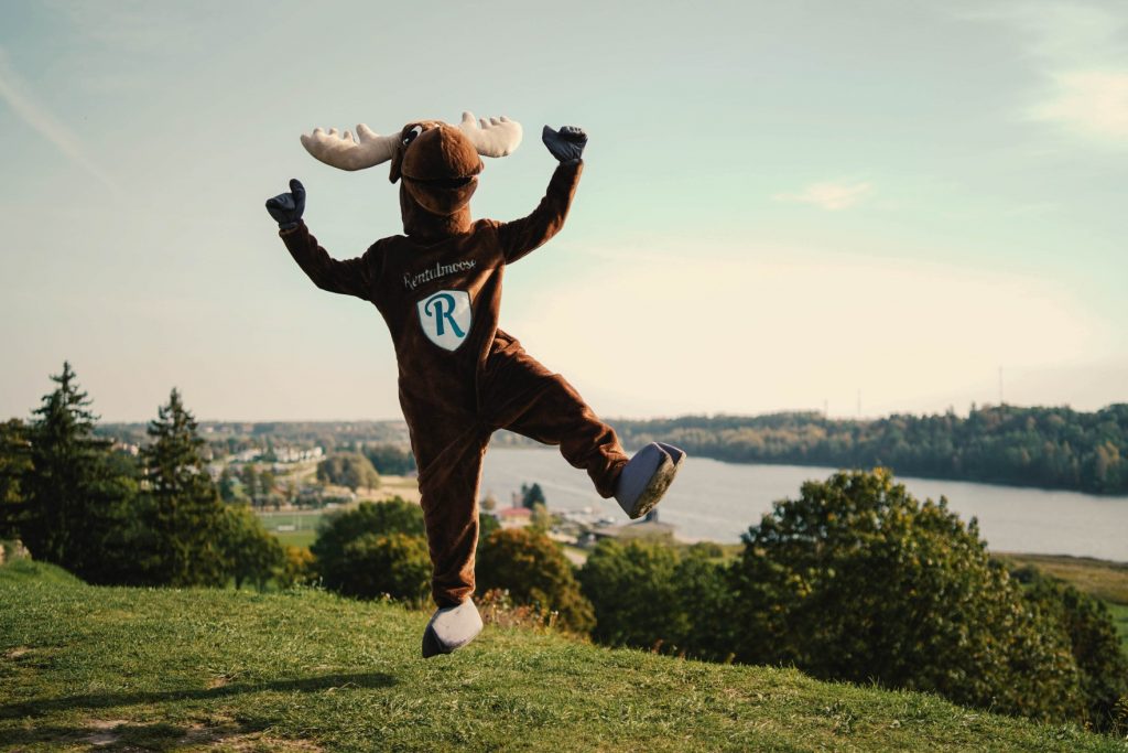 Rental Moose mascot happily jumping on a green field in front of a river in Estonia. Rentalmoose guides answer the question of when the best time to rent a car is.