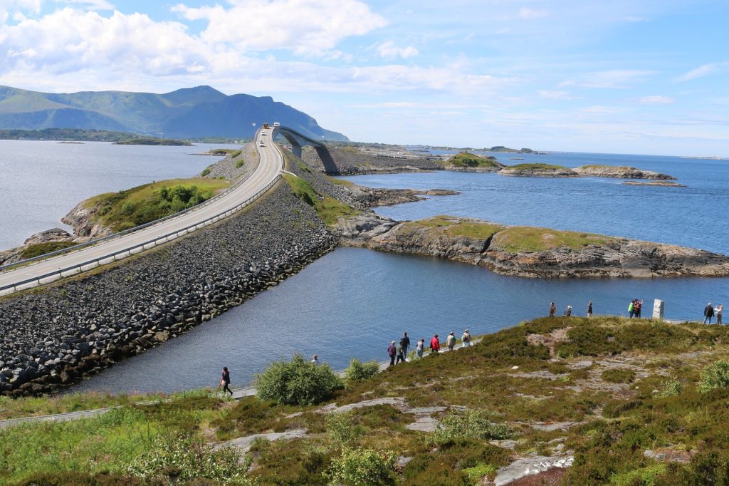 Atlantic Ocean Road in Norway is one of the most scenic drives in the world. Cars crossing a bridge over the ocean, with greenery in the distance and some tourists on the coast in the foreground.